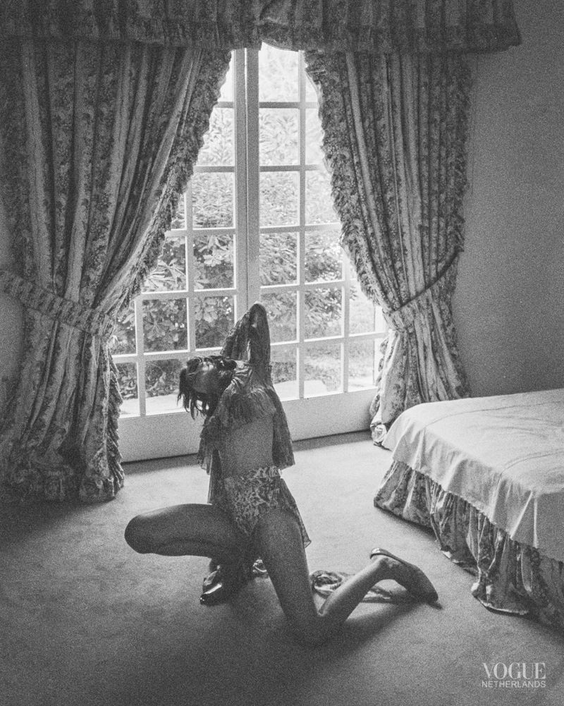 Black and white photograph of a woman kneeling in front of a window with patterned curtains, with a bed visible in the background.