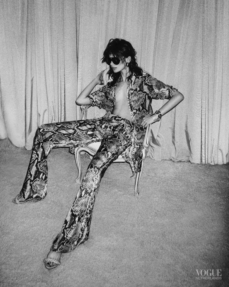 A woman in patterned clothing sits in a chair in a photo studio, wearing sunglasses. The shot is in black and white with a vintage feel.