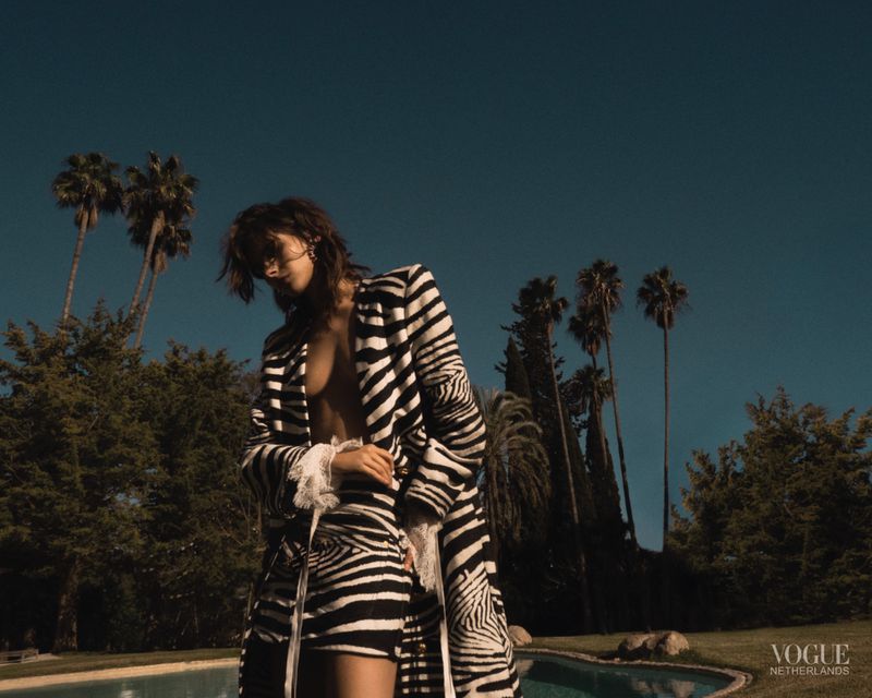 Woman in zebra-striped coat and skirt poses outdoors near a pool against a backdrop of trees and a clear blue sky in a fashion editorial.