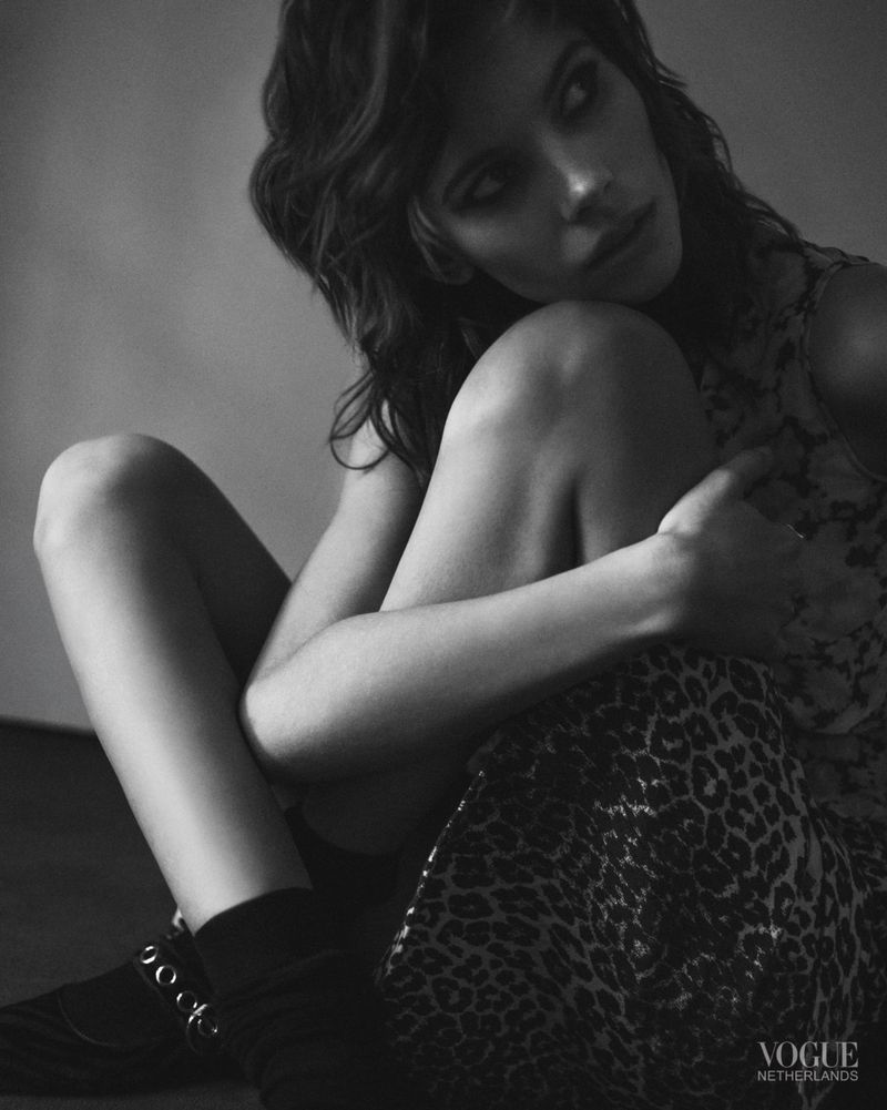 Monochrome portrait of a young woman with curly hair, wearing a patterned dress and ankle boots, sitting indoors against a plain wall.