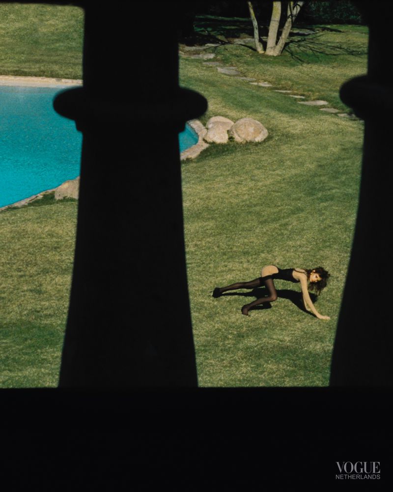 A woman in a black bodysuit and tights lies on a green lawn next to a blue pond, viewed through decorative black bars.