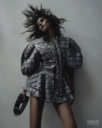 A woman in stylish striped clothing poses indoors with tousled hair, holding a tambourine, in a fashion editorial shot.
