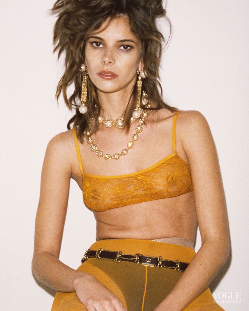 A woman with tousled hair poses in a lace bra top, pants, and jewelry against a white background in a fashion editorial shot.