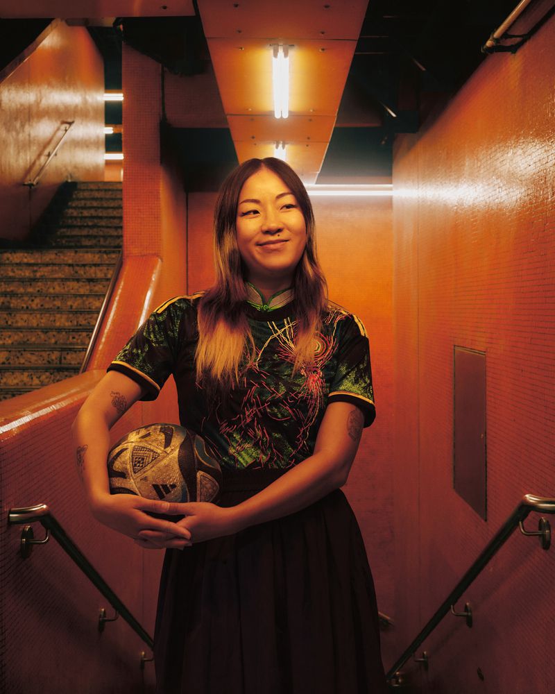 A young adult woman with a soccer ball smiles in an orange-toned stairwell with overhead fluorescent lighting and a tiled wall backdrop.