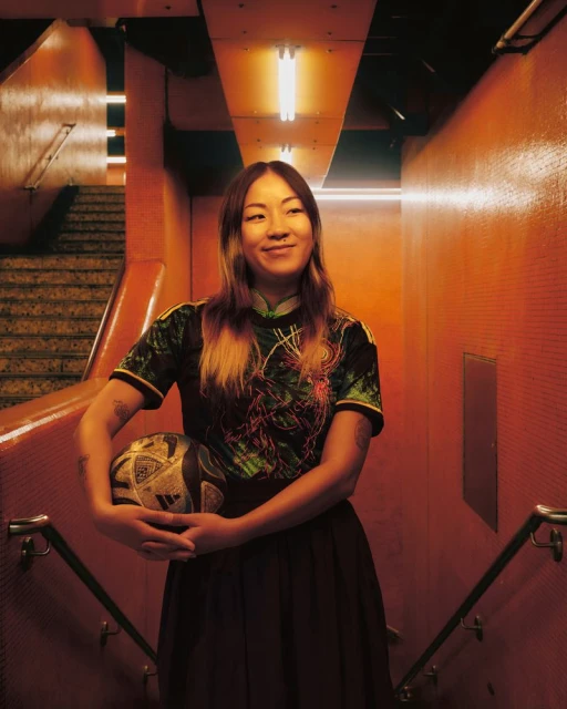 A young adult woman with a soccer ball smiles in an orange-toned stairwell with overhead fluorescent lighting and a tiled wall backdrop.