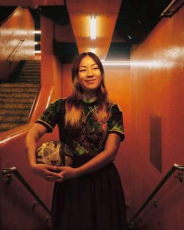 A young adult woman with a soccer ball smiles in an orange-toned stairwell with overhead fluorescent lighting and a tiled wall backdrop.