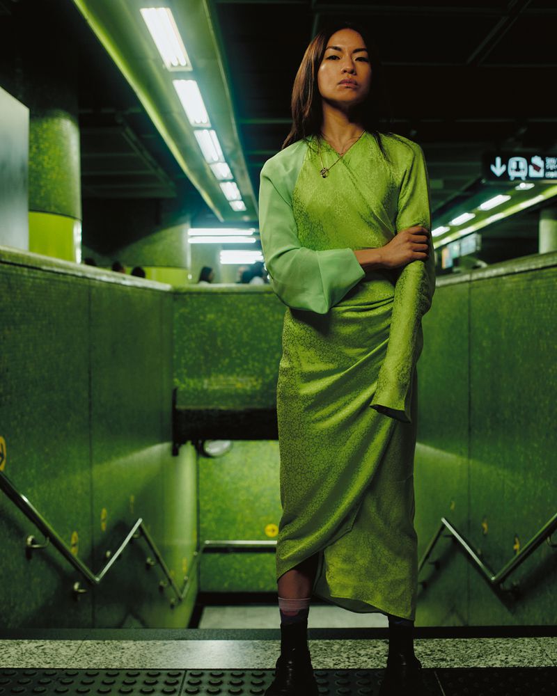 An Asian woman wearing a green wrap dress in a subway, standing on the stairs with her arms crossed in a medium eye-level shot.