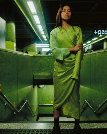 An Asian woman wearing a green wrap dress in a subway, standing on the stairs with her arms crossed in a medium eye-level shot.