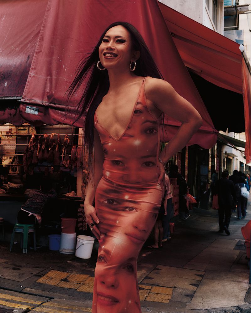 Smiling person in a dress with faces stands outdoors on a city street with market stalls under red awnings, captured in a medium shot.