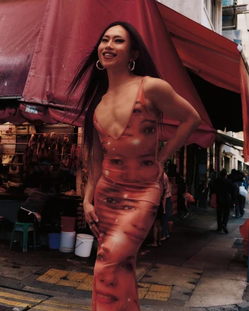 Smiling person in a dress with faces stands outdoors on a city street with market stalls under red awnings, captured in a medium shot.