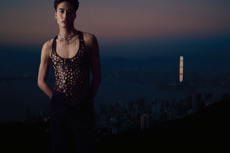 Portrait of an adult male in a black mesh top with a city skyline at night in the background. Fashion, style, and urban photography.