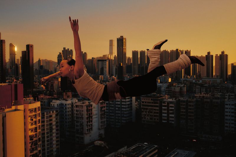 Adult woman with arms raised in an aerial dance pose against an orange sky, wearing black leggings, arm warmers, and a cityscape backdrop.