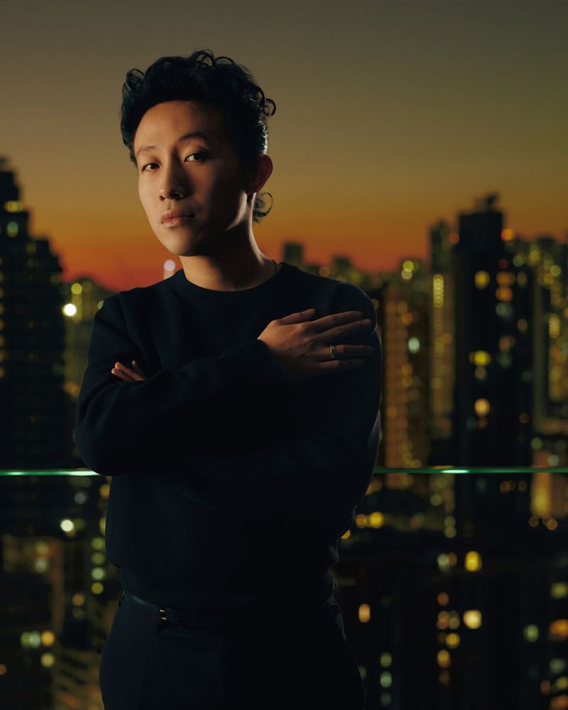 Portrait of a young adult with arms crossed in front of a cityscape at night with warm lighting.