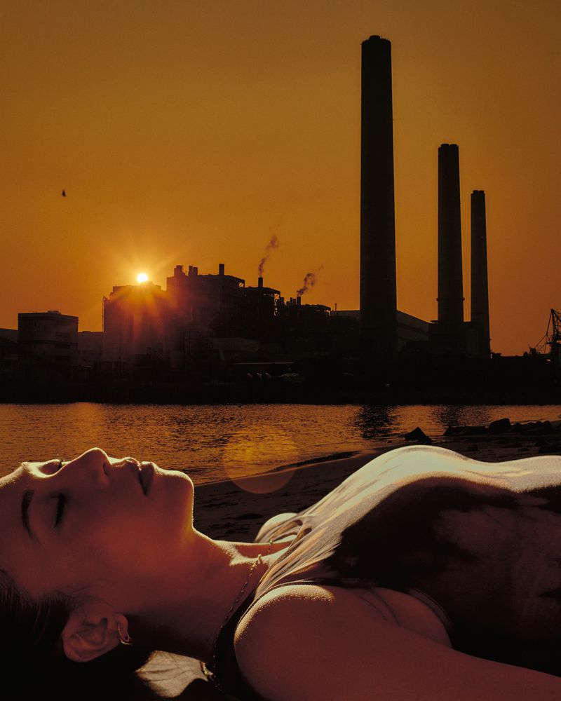 Woman lying down on a beach with eyes closed, illuminated by golden sunset light, with a silhouetted industrial skyline in the background.