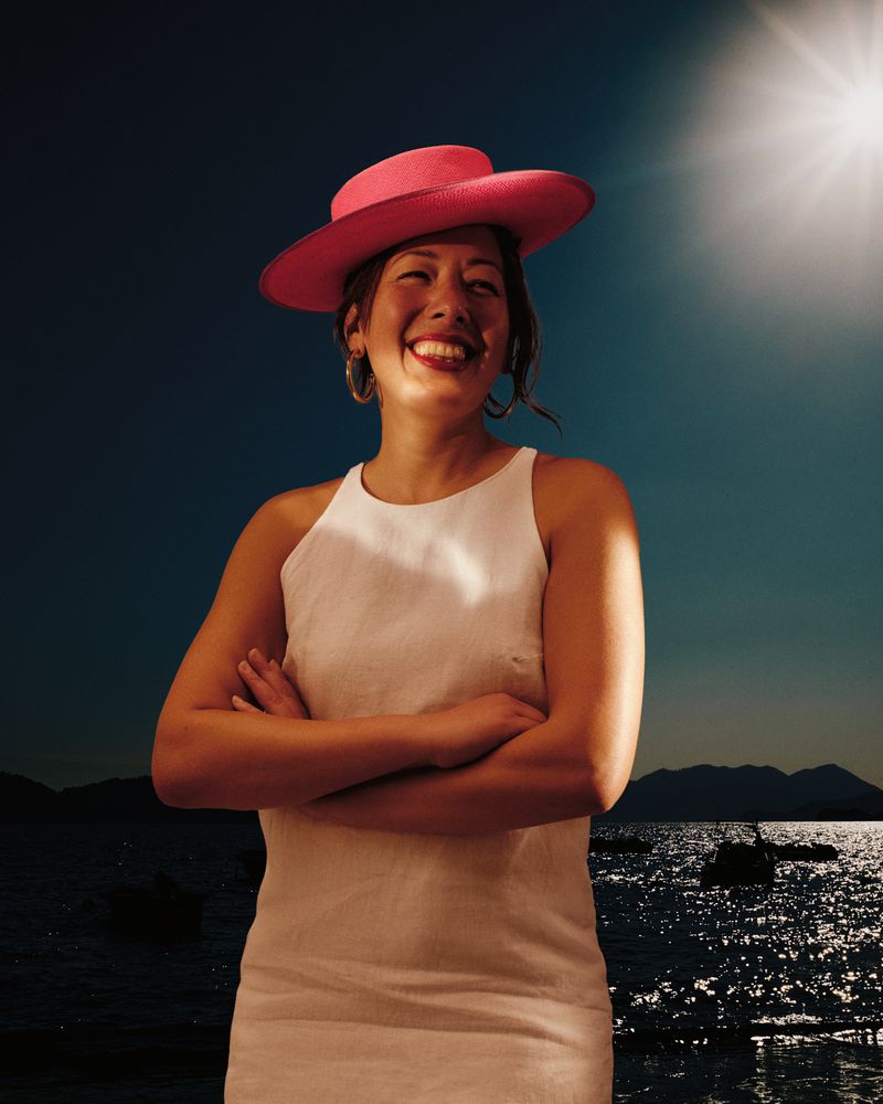 Smiling woman in a white dress and pink hat poses outdoors with arms crossed in front of a seascape with sun glare.