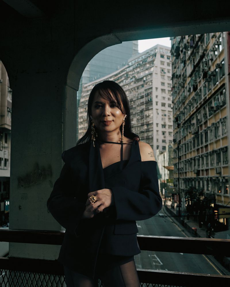 A woman poses wearing a dark blazer in front of tall apartment buildings in a city environment, while standing on a bridge.