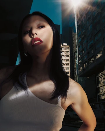 Portrait of an adult woman with dark hair, wearing a white top, with bright red lipstick, posing against an urban building backdrop.