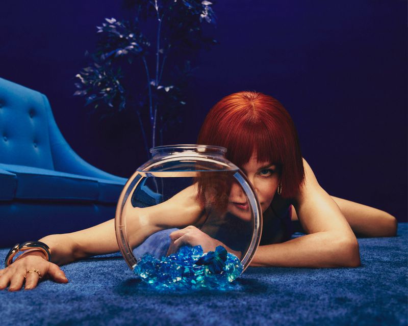 Woman with bob haircut lies on a blue carpet next to a fishbowl filled with blue gems in a modern, moody, indoor studio setting.