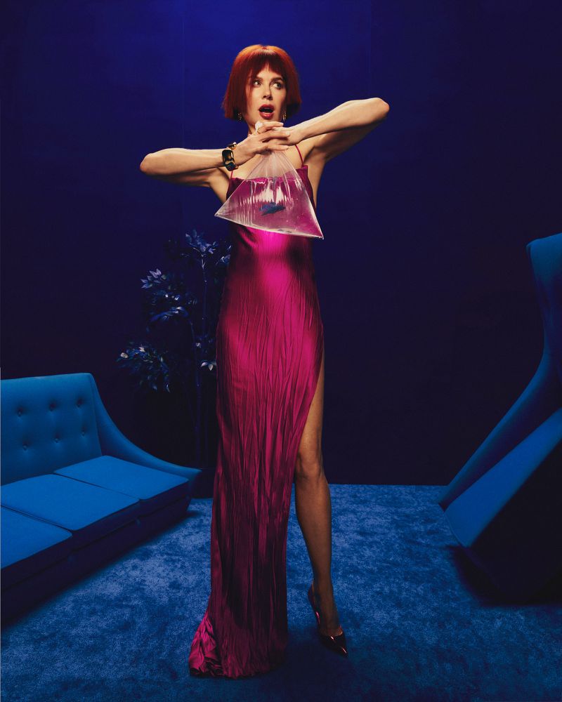 Woman in magenta dress holding a clear plastic bag with water and fish, standing against a blue background with furniture.
