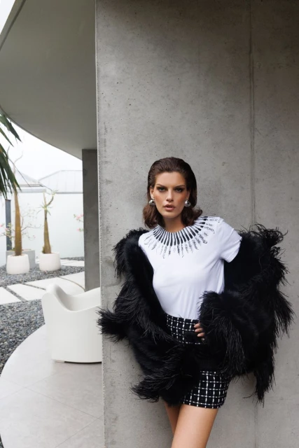A woman poses in a white tee, tweed shorts, and feathered jacket in front of a modern concrete building on a sunny day.