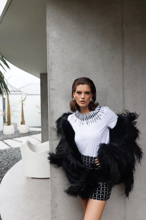 A woman poses in a white tee, tweed shorts, and feathered jacket in front of a modern concrete building on a sunny day.