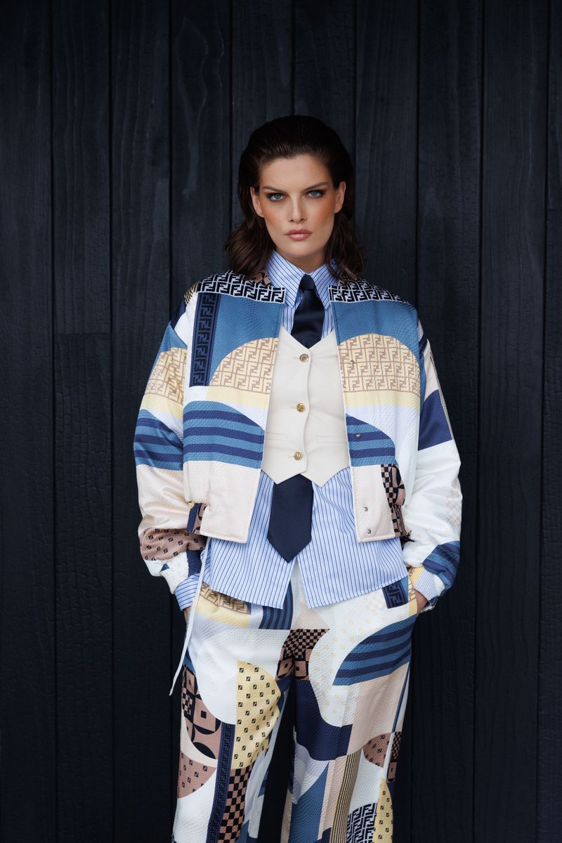 A woman in a patterned jacket, vest, shirt, and pants stands against a dark wood wall, creating a fashion-forward, visually striking image.