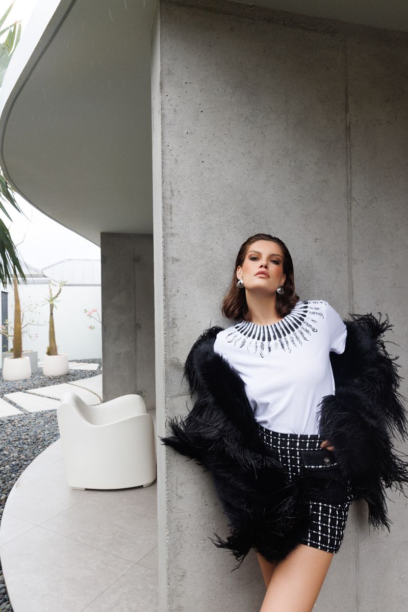 Fashionable adult female in black and white tweed shorts suit with feather bolero posing near modern architectural exterior.