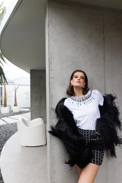 Fashionable adult female in black and white tweed shorts suit with feather bolero posing near modern architectural exterior.