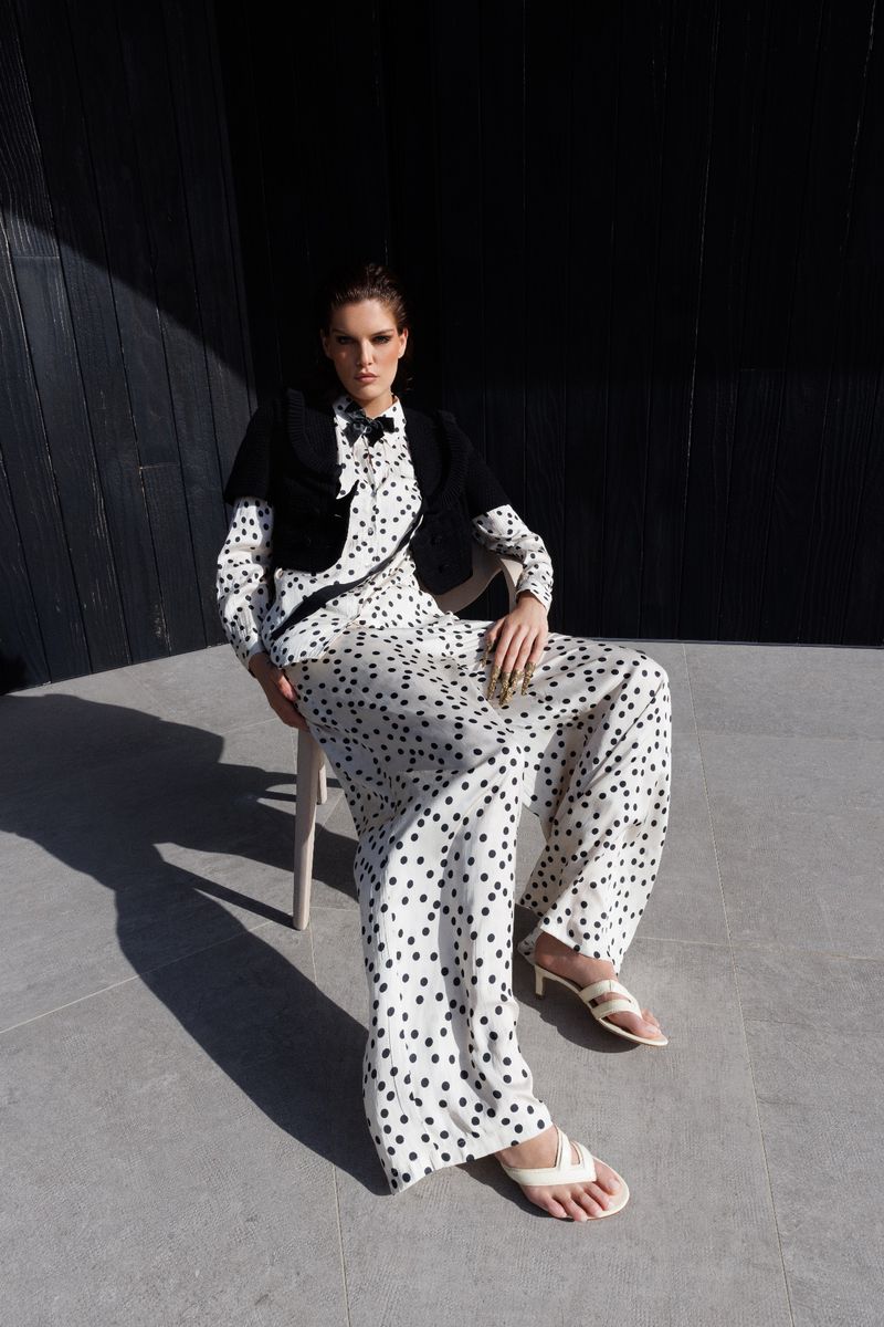 Elegant adult woman in a polka-dot jumpsuit and short bolero jacket poses seated on a chair against a dark wall outside.
