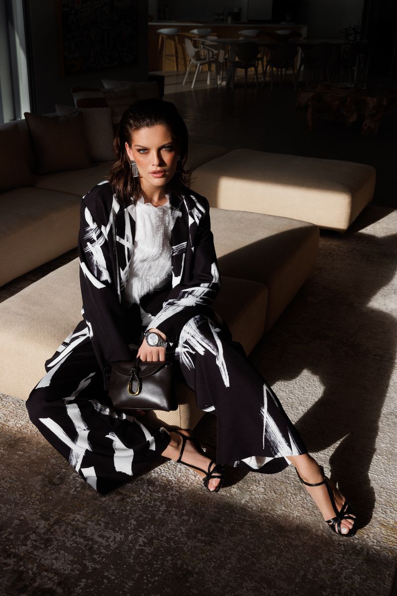 Elegant woman in a black and white printed suit poses indoors with a handbag, jewelry, and stylish heeled sandals, bathed in soft light.