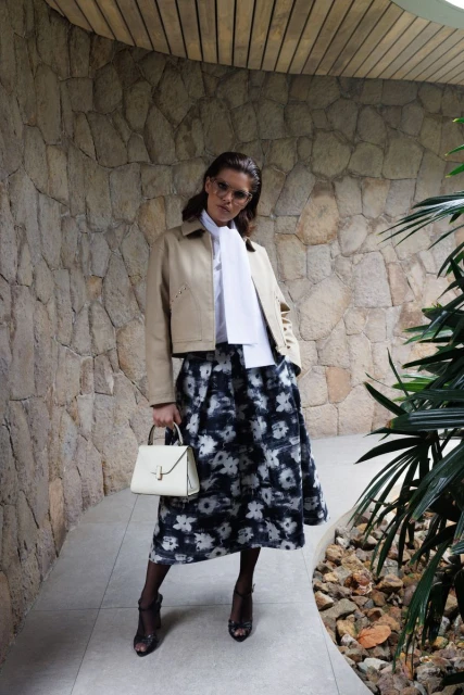 A fashionable woman poses outdoors in a beige jacket, white top, and floral skirt, holding a white handbag, with a textured stone wall backdrop.