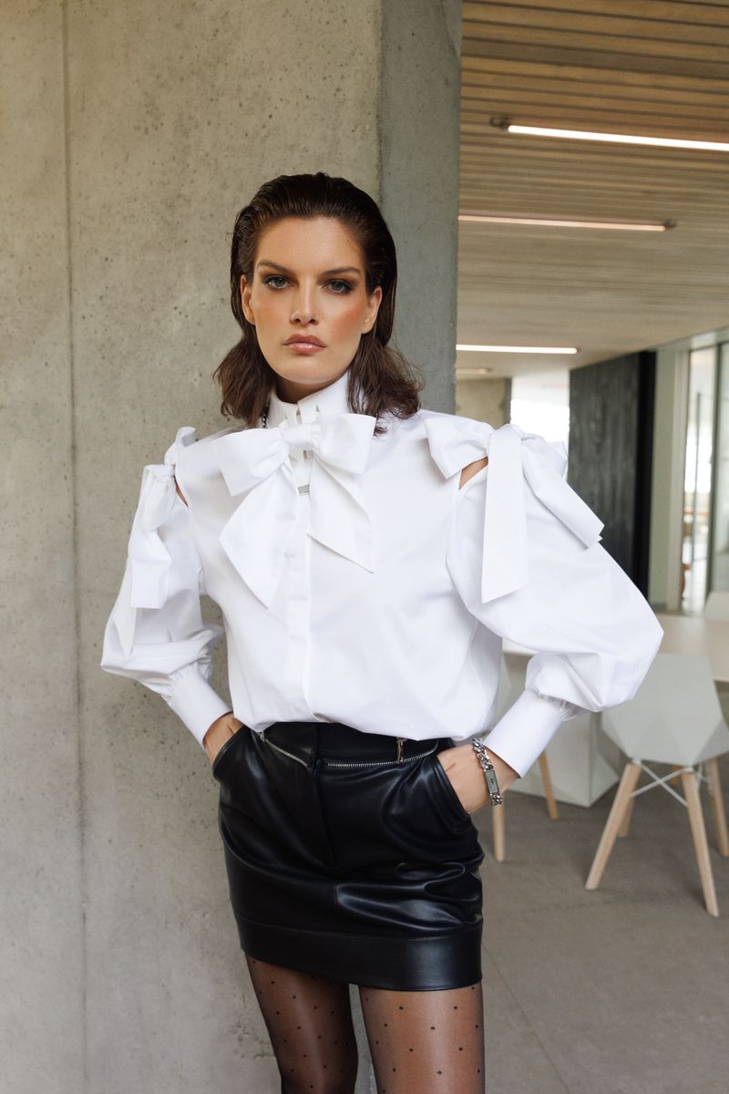 Fashionable brunette woman in a white blouse with bow details and a black mini skirt, posing indoors with a neutral backdrop.