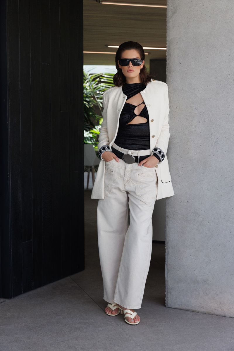 A fashionable adult woman stands in front of a modern doorway wearing a blazer, black top, and oversized light-colored pants with sunglasses.