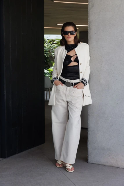 A fashionable adult woman stands in front of a modern doorway wearing a blazer, black top, and oversized light-colored pants with sunglasses.