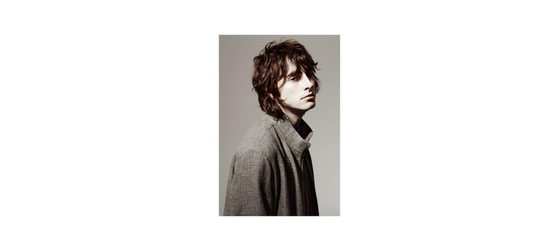Portrait of a brunette adult male in a gray jacket against a light gray studio backdrop, captured in a side view with soft lighting.