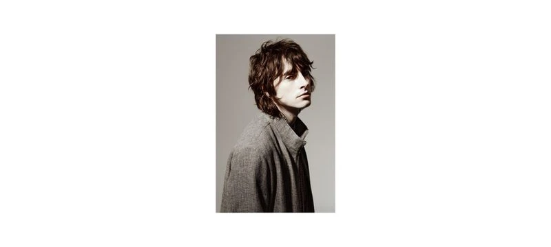 Portrait of a brunette adult male in a gray jacket against a light gray studio backdrop, captured in a side view with soft lighting.