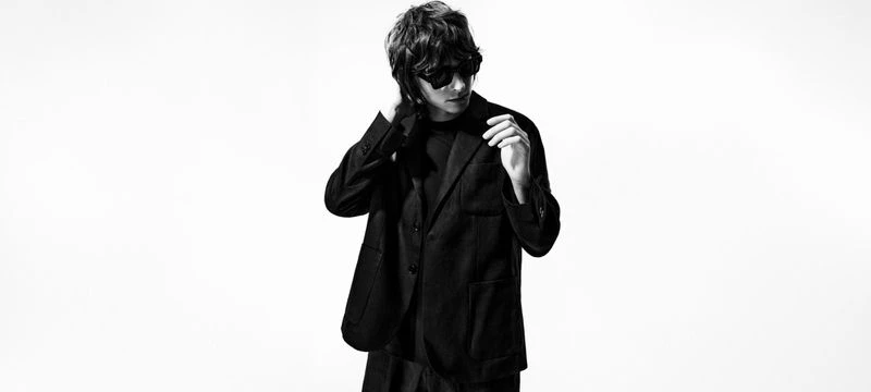 Black-and-white portrait of an adult male with dark hair, wearing sunglasses and a blazer, set against a bright white background.