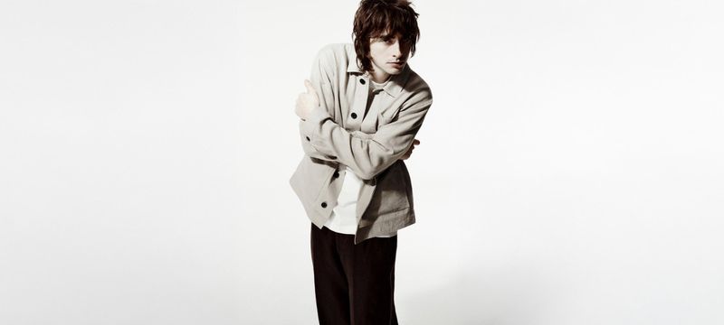 A young adult male poses indoors against a bright white background, wearing a jacket and dark trousers in a medium shot.