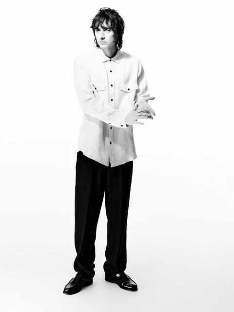 Full-length studio shot of a male model with dark hair, wearing a white button-down shirt and black trousers, standing against a white backdrop.