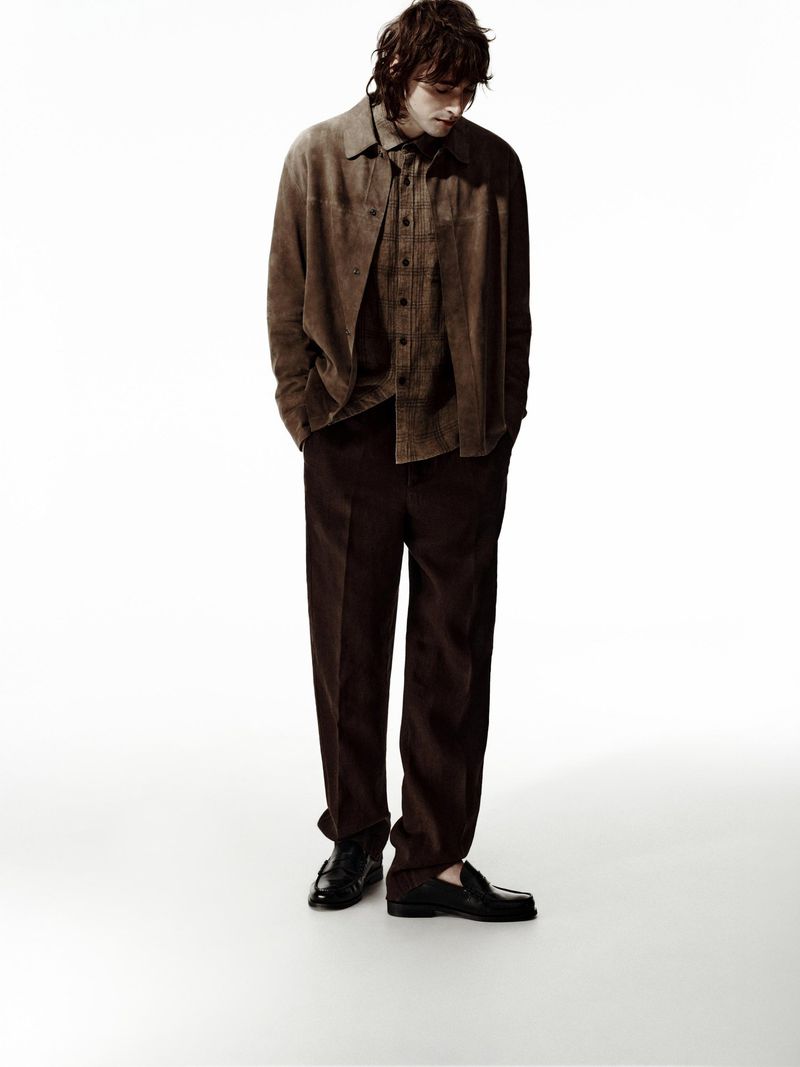 Full-length studio shot of an adult male model in a brown suede shirt, brown pants, and black shoes on a white background looking downward.