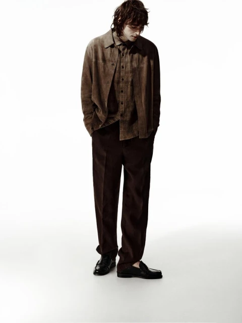 Full-length studio shot of an adult male model in a brown suede shirt, brown pants, and black shoes on a white background looking downward.