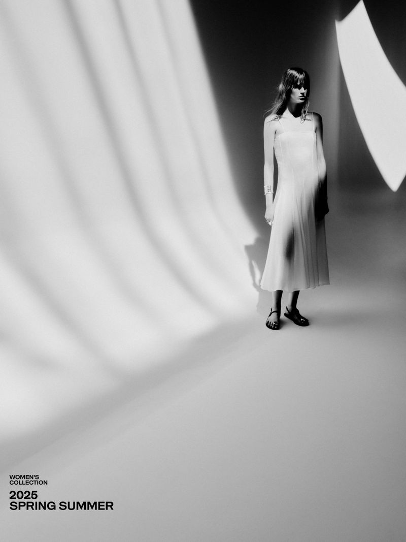 A woman models a long white dress in a studio with dramatic lighting. The image features a minimalistic fashion style, high contrast and monochrome tones.