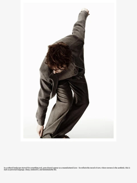 Male fashion model in gray suit coat and trousers poses dynamically against white background in full shot, fashion editorial style.