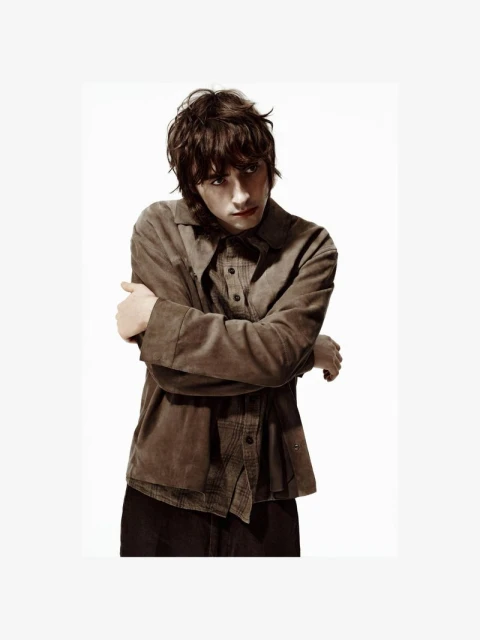 A young adult male poses indoors wearing a brown button-down shirt and dark trousers against a white background in a studio shot.