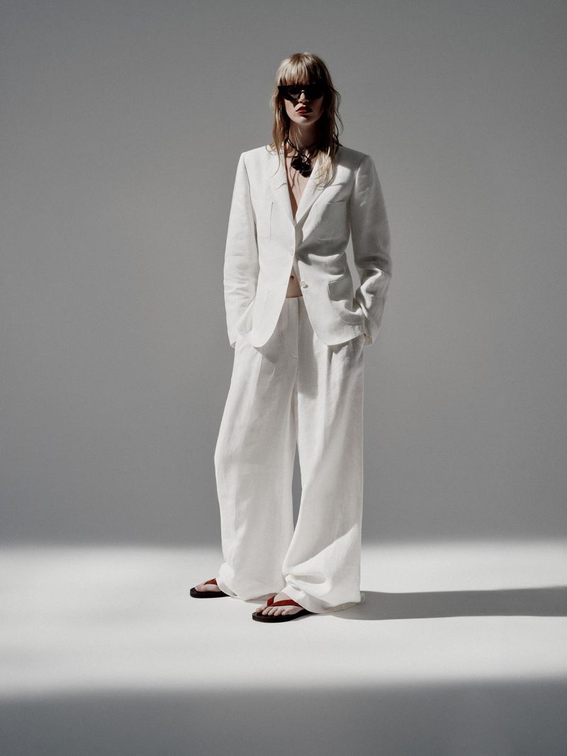 An adult female poses in a white linen suit with wide-leg trousers, a blazer, sunglasses, and sandals, set against a solid gray backdrop.