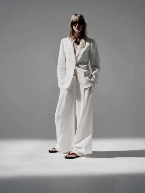 An adult female poses in a white linen suit with wide-leg trousers, a blazer, sunglasses, and sandals, set against a solid gray backdrop.