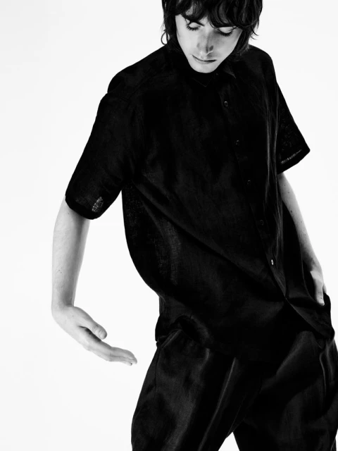 Black and white portrait of a person in a short-sleeved linen shirt and loose pants against a bright backdrop in a studio setting.