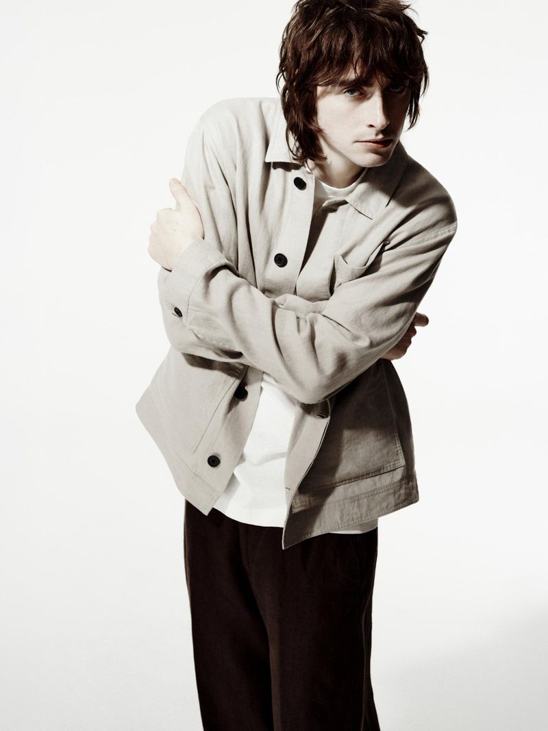 Man in beige jacket and black pants with arms crossed, posing in a full shot against a white backdrop. Fashion editorial style.