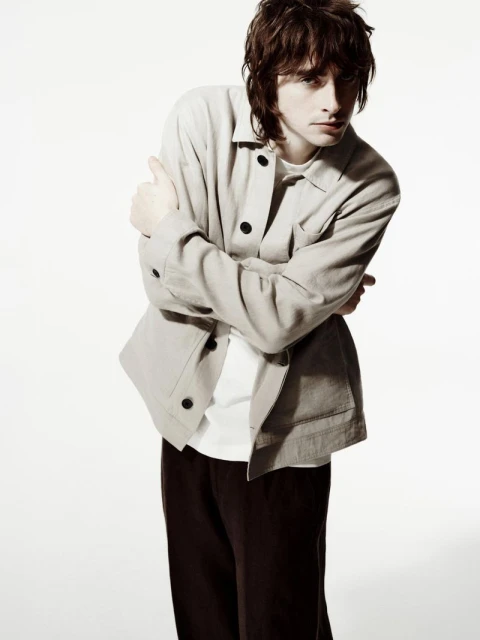 Man in beige jacket and black pants with arms crossed, posing in a full shot against a white backdrop. Fashion editorial style.