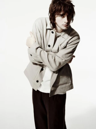 Man in beige jacket and black pants with arms crossed, posing in a full shot against a white backdrop. Fashion editorial style.